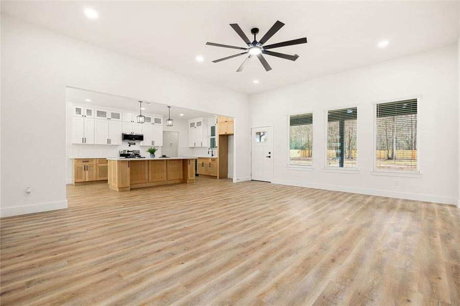 This spacious open-concept living area features modern wood flooring, large windows offering plenty of natural light, and a sleek kitchen with an island and white cabinetry. A stylish ceiling fan adds a contemporary touch.