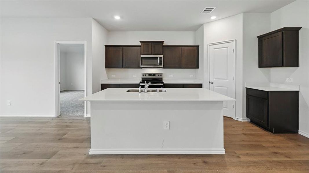 Kitchen with dark brown cabinetry, stainless steel appliances, light stone counters, an island with sink, and light wood finished floors
