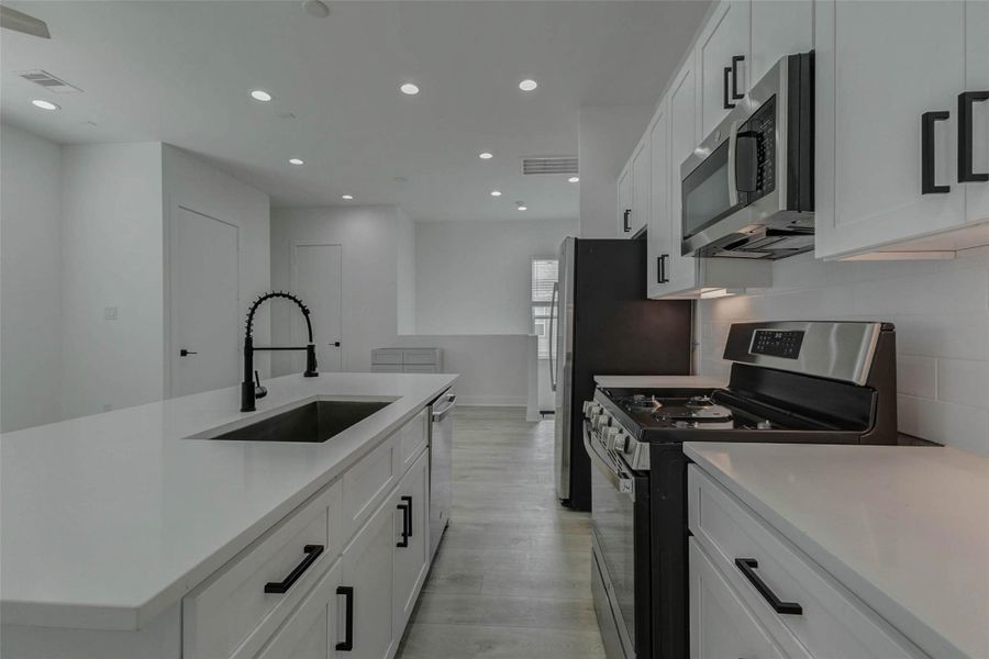 Kitchen close-up featuring a large island sink, matte black pull-down faucet, stainless gas range, built-in microwave, refrigerator, white shaker-style cabinetry, black hardware, and bright under-cabinet lighting. The clean finishes and open sightline create a crisp, functional, modern cooking space.