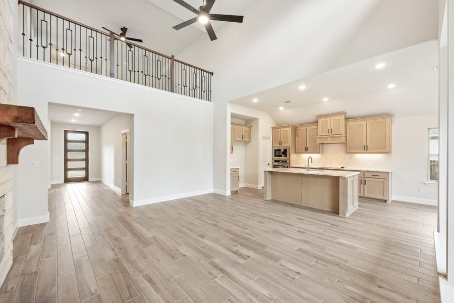 Open-concept living area with high ceilings, kitchen island, light wood floors, and second-floor balcony railing