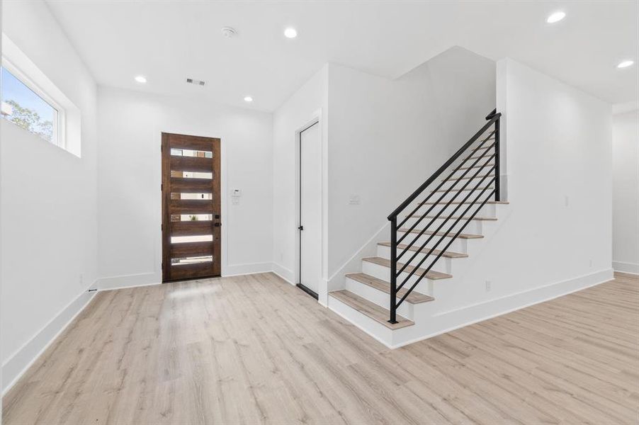 Entryway with bright lighting, featuring a wooden door with glass panels, a sleek black staircase with wooden steps, and luxury vinyl wood flooring. The white walls enhance the space's clean and spacious feel.