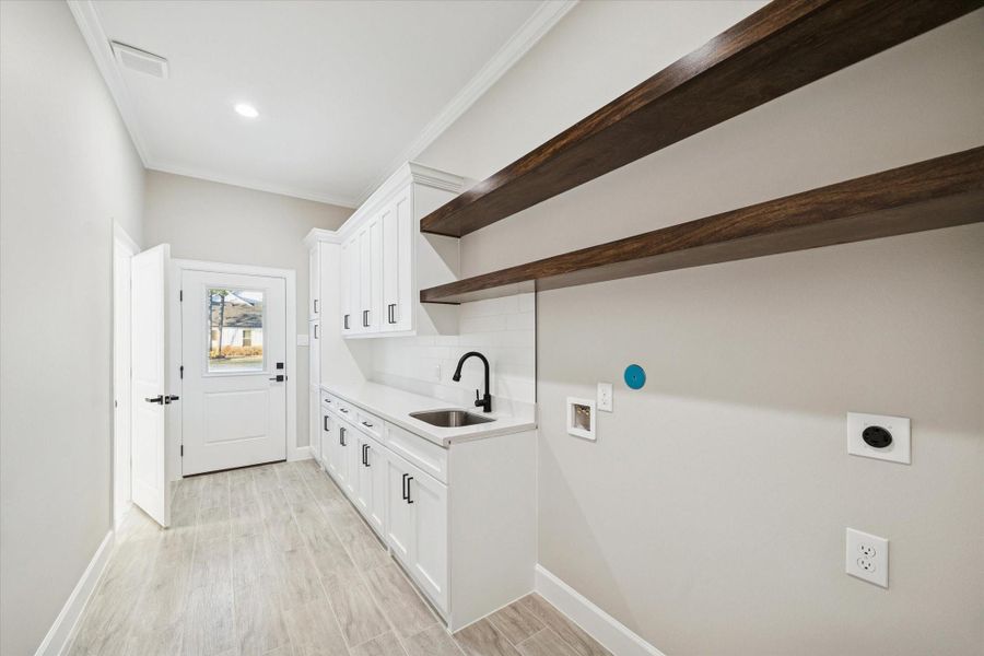Utility room with upper and lower closed storage.  Open shelving above washer and dryer space, utility sink.Half-light exterior door with inner shade leads to side of the home and driveway.  Door on the left leads to the primary bath.