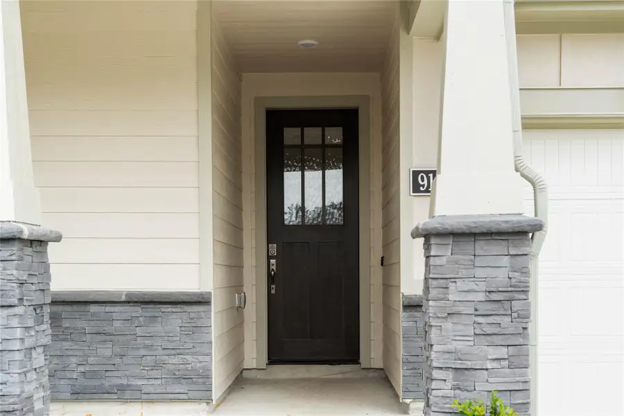 Welcome to a charming, well-maintained front porch, perfect for relaxing and enjoying the outdoors. The porch features sophisticated grey stacked stone accents around the base of the columns and the house's foundation, complemented by neutral beige siding and smooth trim.