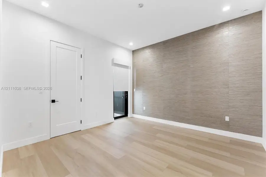 Interior space featuring a textured accent wall, light-toned wood-finish flooring, recessed ceiling lighting, a solid-core interior door with matte black hardware, and a window fitted with a roller shade