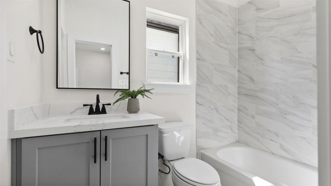 Modern secondary bathroom featuring a faux marble tile, a modern sleek gray custom vanity with a quartz countertop, custom modern framed matte black mirror, and a full-size bathtub surrounded by stone tiles.