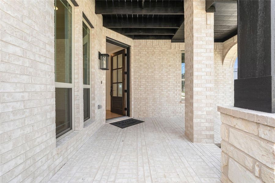 Covered front porch with light brick walls, dark wood beams, and open glass-panel entry door Covered front porch with light brick walls, dark wood beams, and open glass-panel entry door