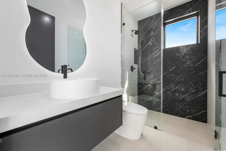 Floating vanity with a stone-composite countertop, vessel sink, backlit organic-shaped mirror, and walk-in shower featuring dark-toned tiled walls and a frameless glass enclosure