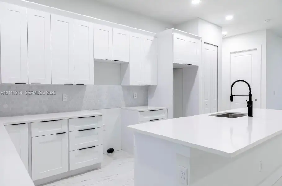 Kitchen featuring white shaker-style cabinetry, a central island with an undermount sink, a matte black gooseneck faucet, light-toned stone-finish countertops, and a hexagonal tile backsplash Kitchen featuring white shaker-style cabinetry, a central island with an undermount sink, a matte black gooseneck faucet, light-toned stone-finish countertops, and a hexagonal tile backsplash
