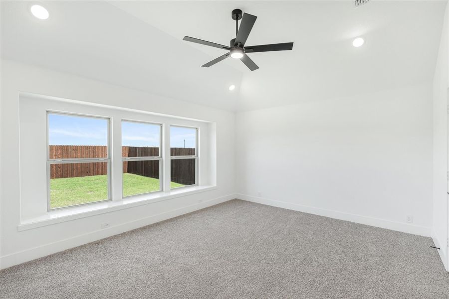 Empty bedroom with gray carpet, black ceiling fan, and triple windows overlooking backyard