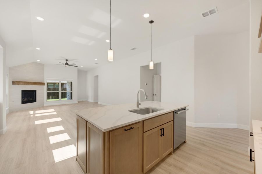 Kitchen island with sink, wood cabinets, and pendant lights open to living room with fireplace and light wood floors