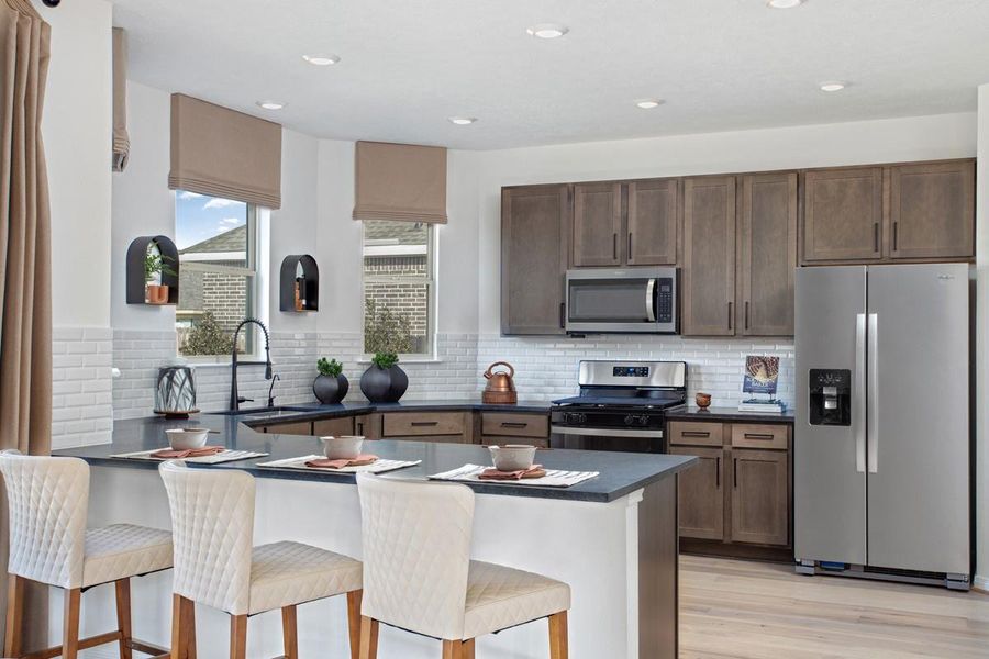 The kitchen exudes warmth with stunning Black Mist granite countertops perfectly paired with the rich, inviting tone of the Woodmont cabinetry in a Smolder stain. This combination creates a truly gorgeous and cohesive aesthetic. Additionally, this kitchen features an extended breakfast bar, stainless steel Whirlpool appliances, gorgeous Daltile backsplash tile, recessed lighting, and large windows for beautiful outdoor lighting.