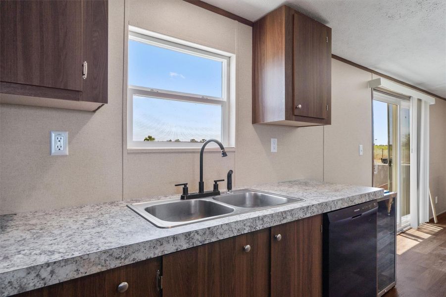 Laminate countertops and wood cabinetry frame a double sink below a window framing blue skies.