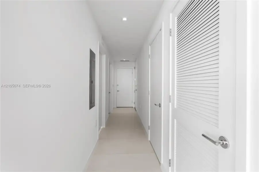 Hallway featuring neutral-toned tile flooring, recessed ceiling lighting, and solid core doors with brushed metal hardware, including a louvered closet door