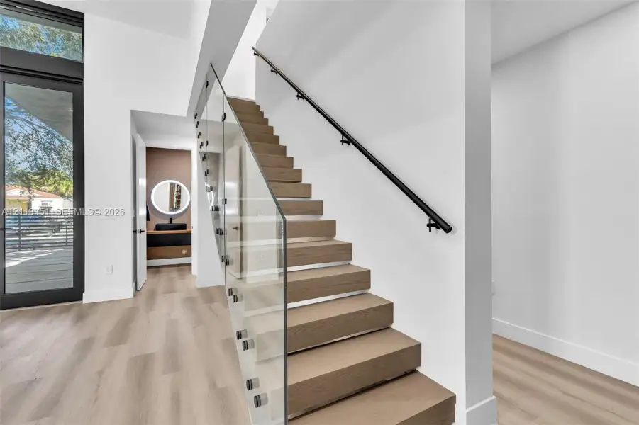 Staircase featuring wood-finish treads, a glass panel railing with metal hardware, and a matte black wall-mounted handrail, complemented by wood-finish flooring Staircase featuring wood-finish treads, a glass panel railing with metal hardware, and a matte black wall-mounted handrail, complemented by wood-finish flooring