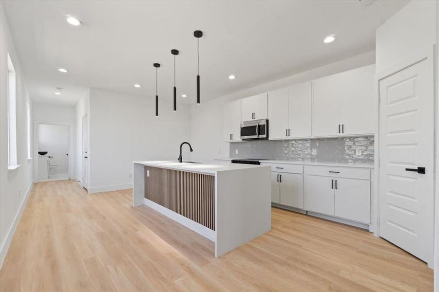 Kitchen featuring tasteful backsplash, light wood-style floors, stainless steel microwave, and an island with sink