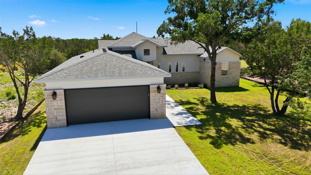 Contemporary residence with a gray shingle roof, stone and stucco exterior, an attached garage with a dark panel door, and mature trees on a landscaped lot Contemporary residence with a gray shingle roof, stone and stucco exterior, an attached garage with a dark panel door, and mature trees on a landscaped lot
