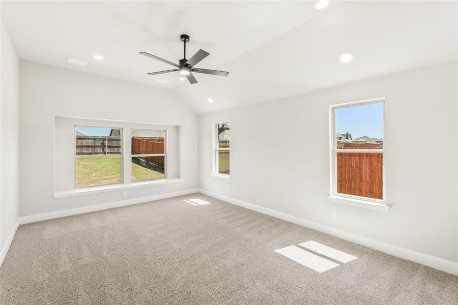 Empty bedroom with vaulted ceiling, ceiling fan, carpet flooring, and multiple windows with backyard views Empty bedroom with vaulted ceiling, ceiling fan, carpet flooring, and multiple windows with backyard views