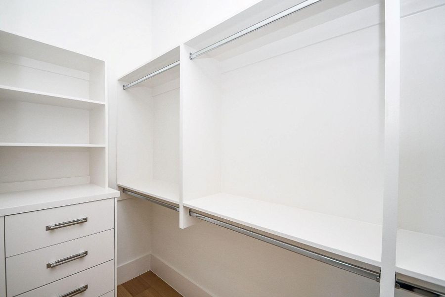 This photo shows a spacious, modern walk-in closet with ample shelving and hanging space. It features sleek white cabinetry and drawers with metal handles, offering plenty of storage options.