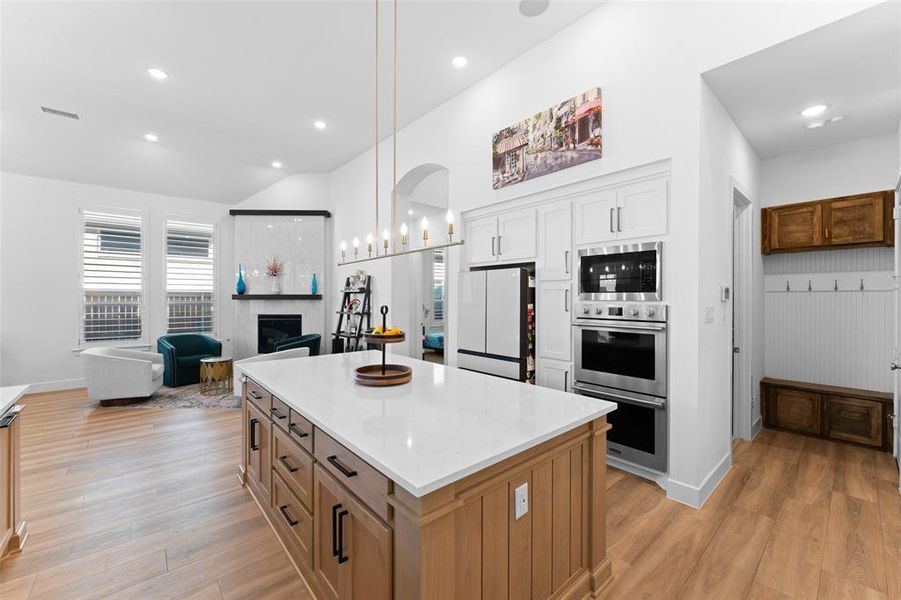 Kitchen with open floor plan, hanging light fixtures, brown cabinetry, a center island, and light wood finished floors Kitchen with open floor plan, hanging light fixtures, brown cabinetry, a center island, and light wood finished floors