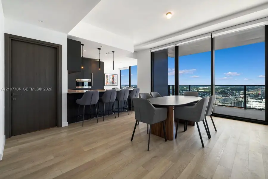 Open-concept interior featuring light wood-finish flooring, floor-to-ceiling windows, matte black cabinetry, built-in stainless steel appliances, and integrated pendant lighting