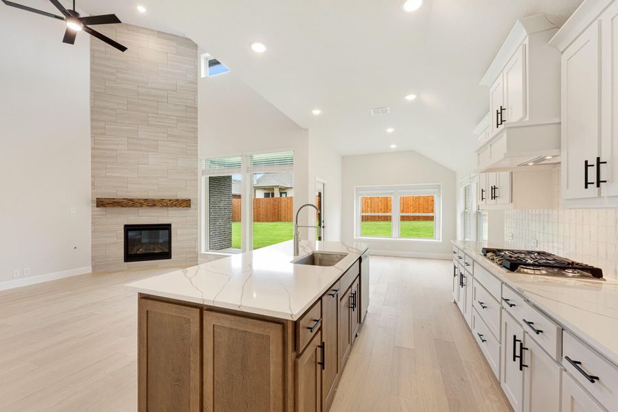 Open kitchen with large island, quartz countertops, white cabinets, and tile fireplace under vaulted ceiling Open kitchen with large island, quartz countertops, white cabinets, and tile fireplace under vaulted ceiling