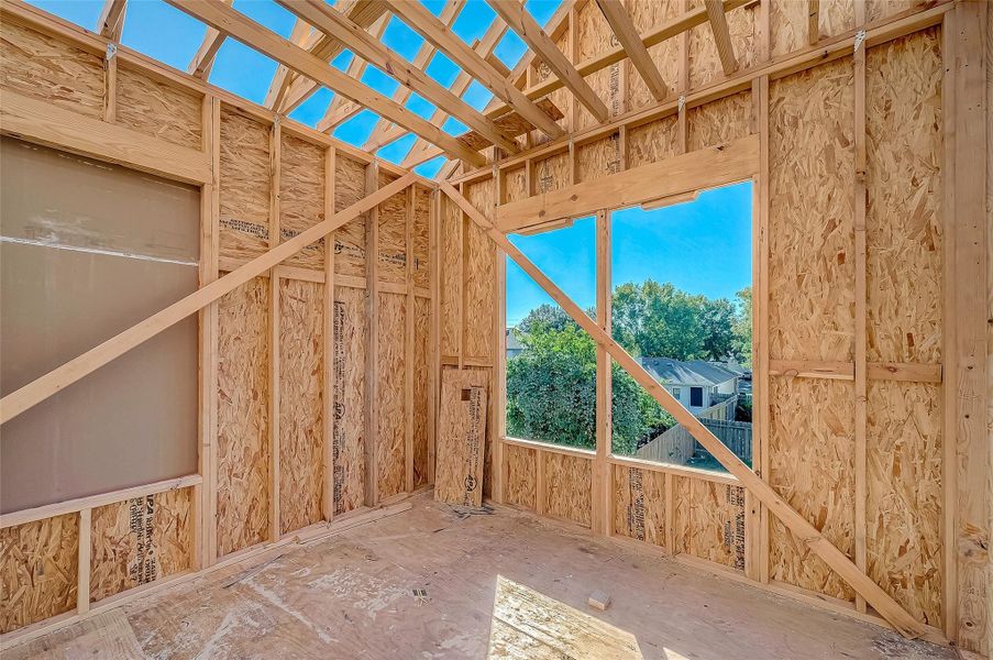 This photo shows a partially constructed room with exposed wooden framing and large window openings, offering a view of trees and neighboring houses. Ideal for customization in a new home build.