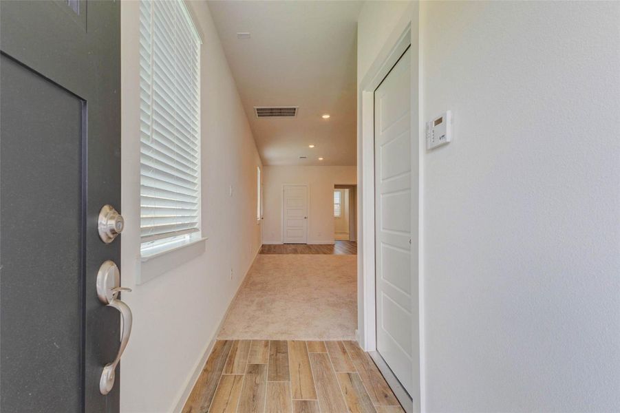 Bright entryway with a black front door, window blinds, wood-look tile flooring, recessed lighting, and a long hallway leading toward the main living areas. The neutral color palette and clean finishes create an inviting first impression with a fresh, modern feel.