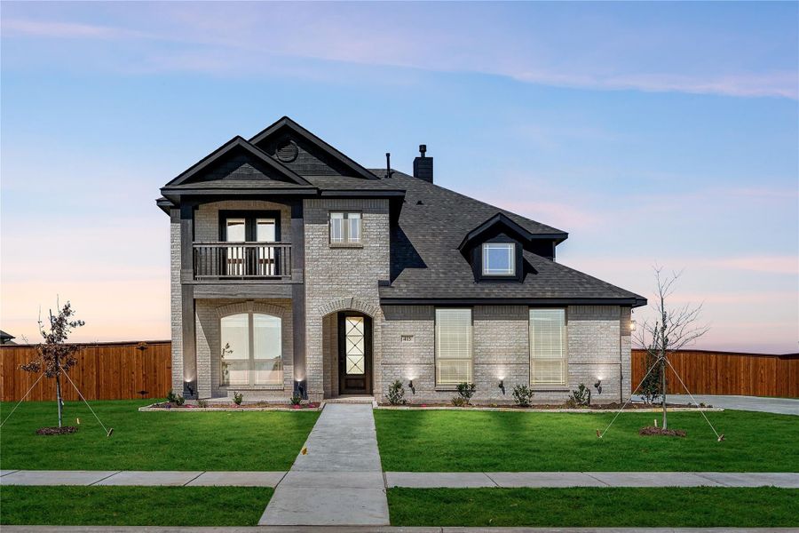 Two-story brick home with arched entry, second-floor balcony, dark roof, and manicured front lawn at dusk Two-story brick home with arched entry, second-floor balcony, dark roof, and manicured front lawn at dusk