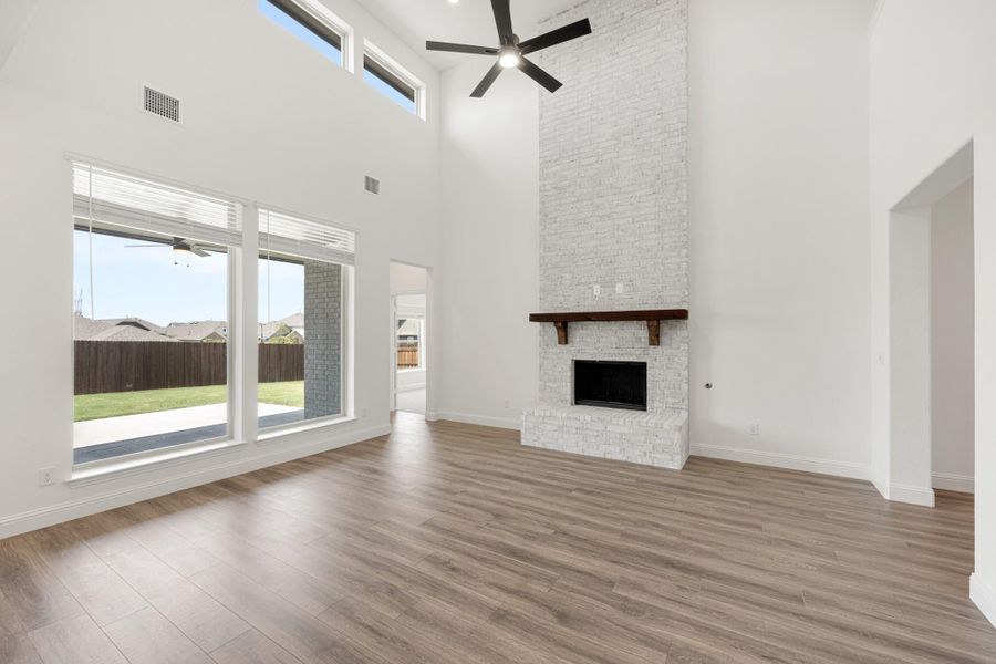 Open living room with two-story ceiling, white brick fireplace, wood floors, and large windows overlooking backyard Open living room with two-story ceiling, white brick fireplace, wood floors, and large windows overlooking backyard