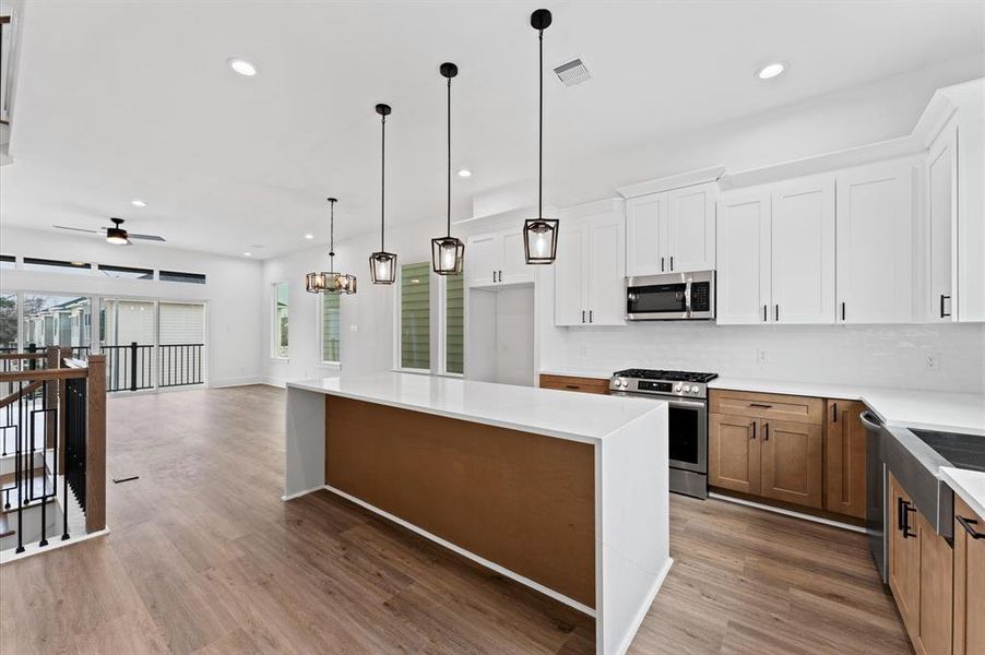 This modern kitchen features a spacious open layout with sleek white cabinetry, contrasting wood accents, and stainless steel appliances. The large island provides ample workspace, and the contemporary pendant lighting adds elegance. The space flows into a bright living area with large windows and wood flooring.