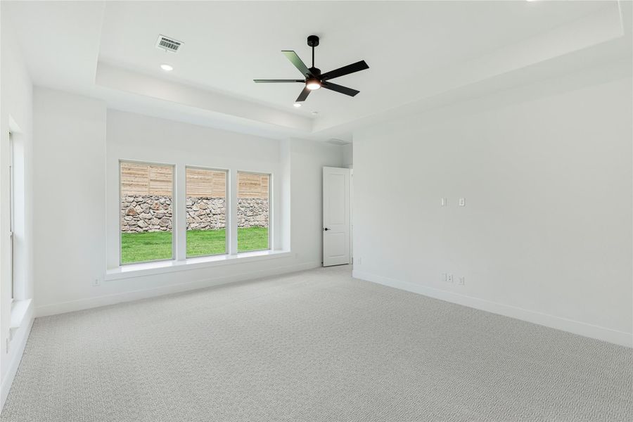 Empty bedroom with tray ceiling, black ceiling fan, carpet flooring, and triple windows overlooking backyard