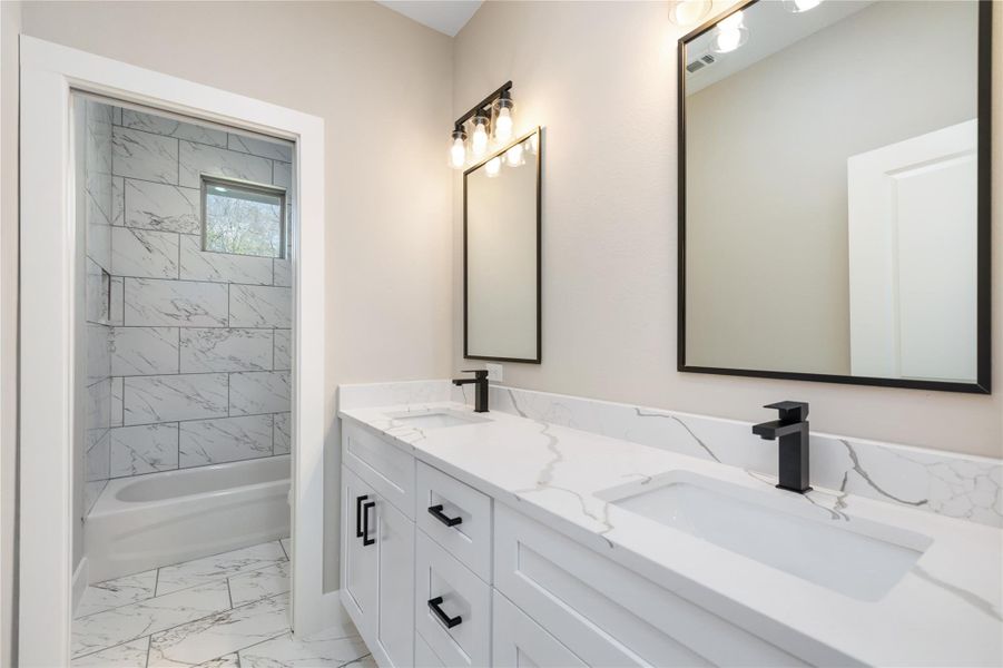 The modern secondary bathroom boasts a stylish dual-sink vanity with sleek black fixtures and large mirrors that enhance the space. A tub-shower combo, adorned with elegant marble-inspired tiles, adds a touch of luxury. Ample natural light floods the room, highlighting the clean, contemporary design and creating a bright, inviting atmosphere.