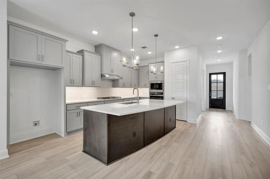 Kitchen featuring hanging lights, a kitchen island with sink, gray cabinetry, light wood-type flooring, and light stone counters Kitchen featuring hanging lights, a kitchen island with sink, gray cabinetry, light wood-type flooring, and light stone counters