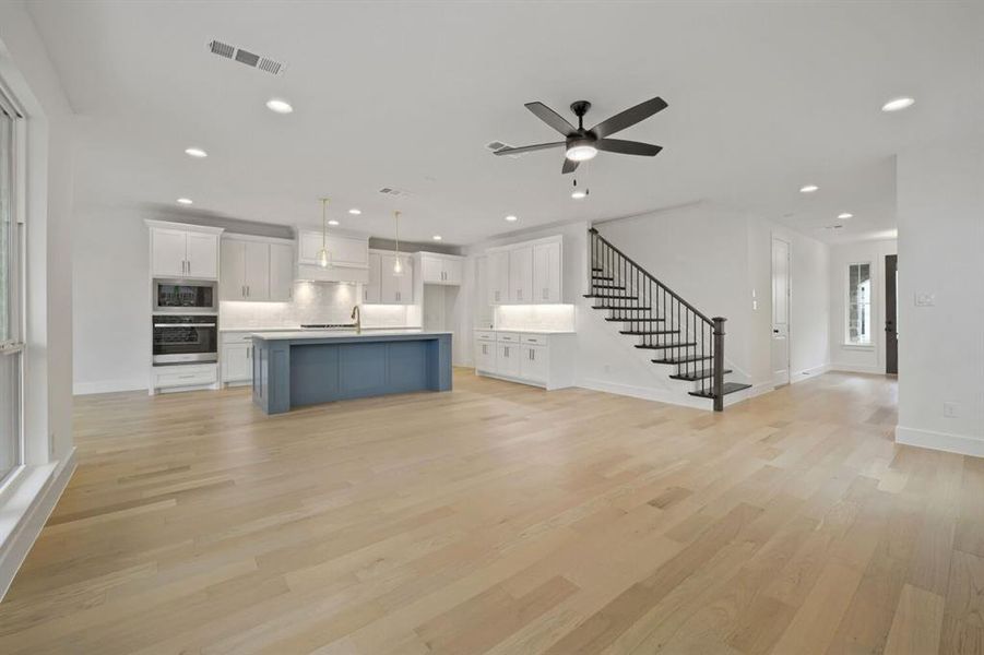 Open-concept living space featuring light wood-finish flooring, a contemporary kitchen with a blue-hued island, white cabinetry, integrated stainless steel appliances, and a staircase with wrought iron balusters Open-concept living space featuring light wood-finish flooring, a contemporary kitchen with a blue-hued island, white cabinetry, integrated stainless steel appliances, and a staircase with wrought iron balusters
