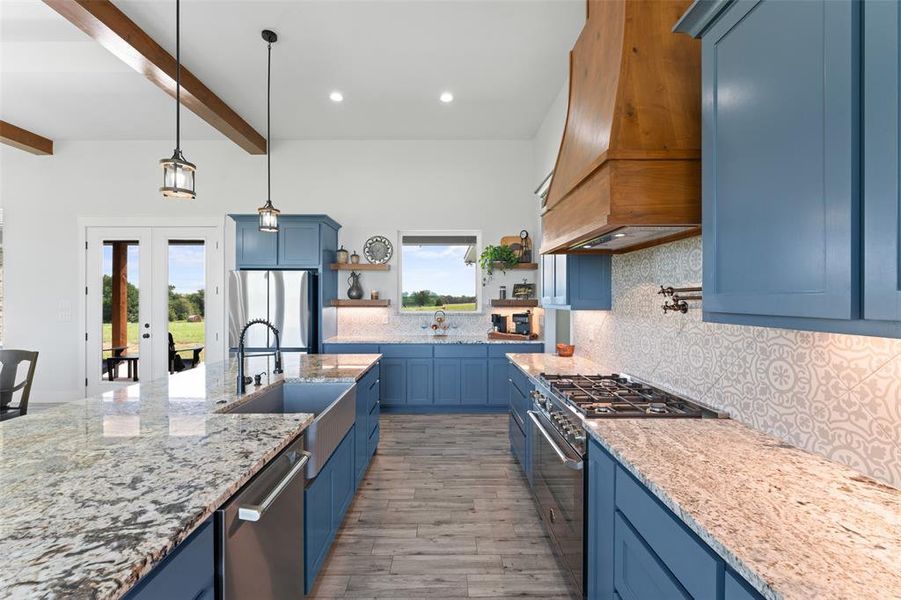 Gourmet kitchen featuring blue cabinetry, granite countertops, a farmhouse sink, stainless steel appliances, and a wood range hood