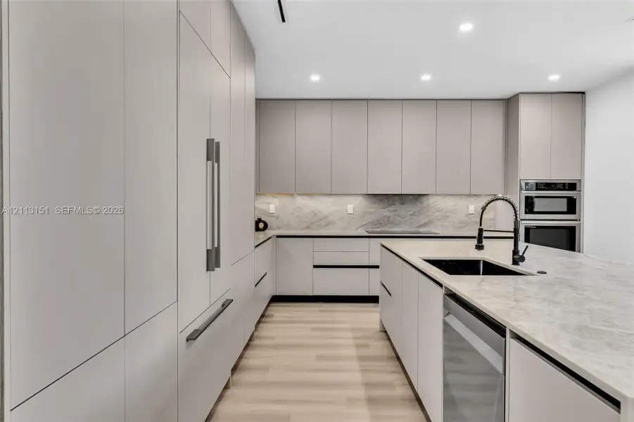 Kitchen featuring flat-panel cabinetry, stone countertops, a waterfall-edge island, built-in stainless steel appliances, and wood-finish flooring Kitchen featuring flat-panel cabinetry, stone countertops, a waterfall-edge island, built-in stainless steel appliances, and wood-finish flooring