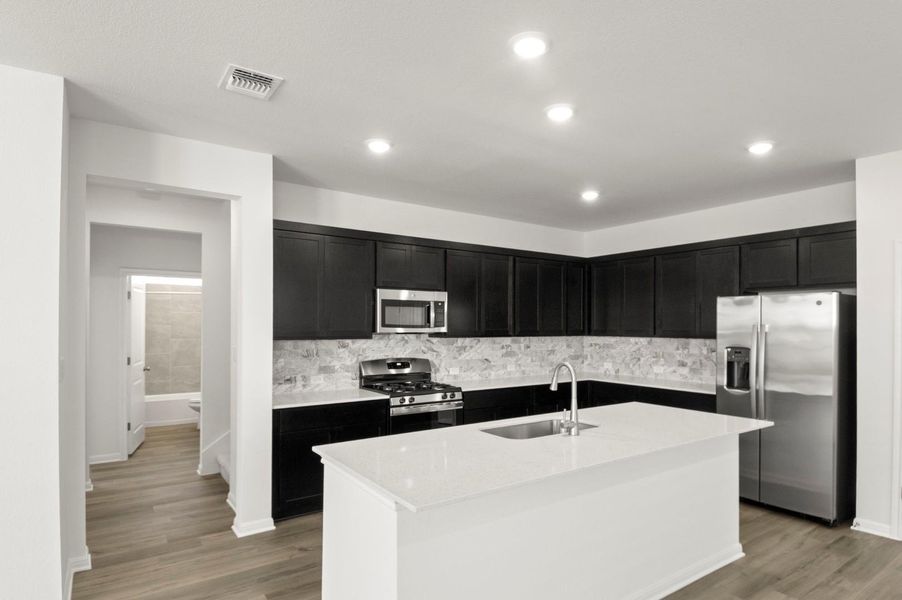 Kitchen with dark cabinets, appliances with stainless steel finishes, decorative backsplash, light stone countertops, and an island with sink