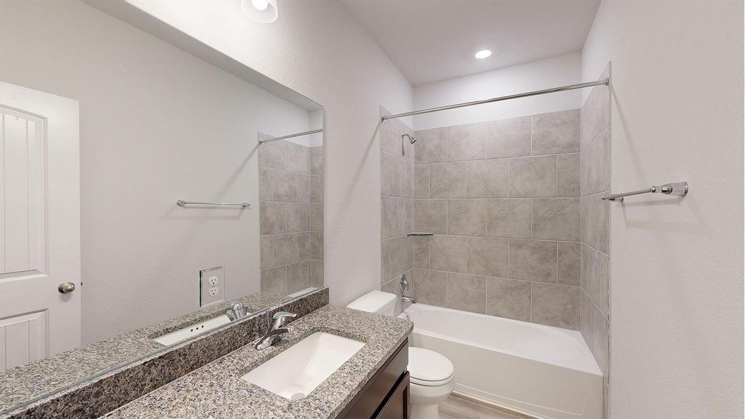 Bathroom featuring a granite-look vanity with an integrated sink, a full-width mirror, a shower-over-tub combination with large format tile surround, and wood-finish flooring Bathroom featuring a granite-look vanity with an integrated sink, a full-width mirror, a shower-over-tub combination with large format tile surround, and wood-finish flooring