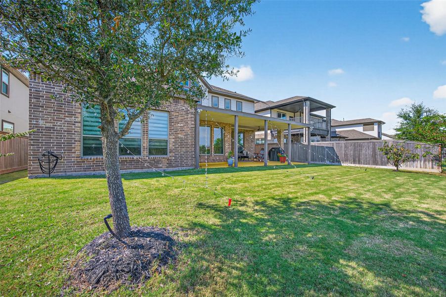 This photo shows a spacious backyard with a well-maintained lawn, a mature tree, and a covered patio attached to a brick house. The space is ideal for outdoor activities and relaxation. This photo shows a spacious backyard with a well-maintained lawn, a mature tree, and a covered patio attached to a brick house. The space is ideal for outdoor activities and relaxation.