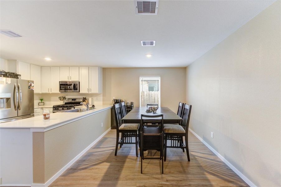 This photo showcases an open-concept kitchen and dining area. The kitchen features modern stainless steel appliances, white cabinetry, and a breakfast bar. The dining area is spacious, with a large table and seating for six, highlighted by natural light from a window. The flooring is a light vinyl plank finish, enhancing the bright and airy feel of the space.