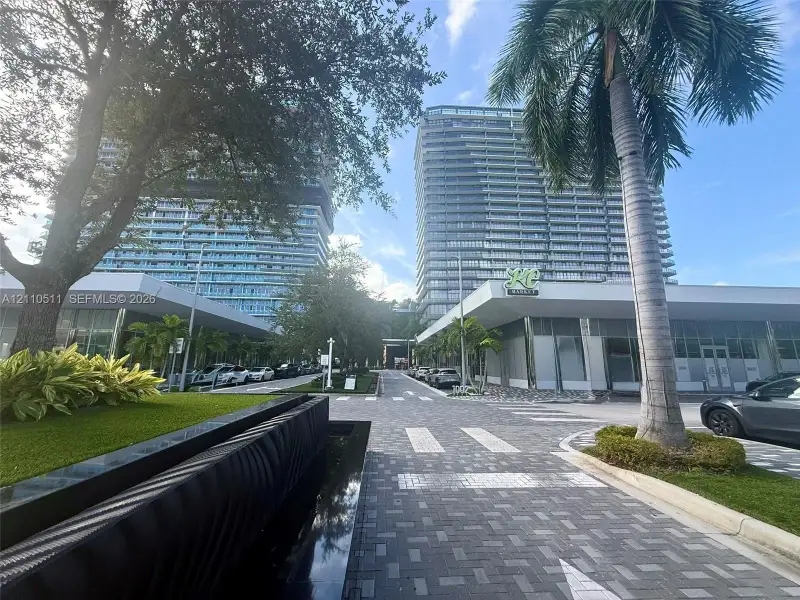 High-rise architectural exterior featuring glass curtain walls, multi-level structural design, stone-paver pedestrian walkways, decorative water feature, and mature palm landscaping