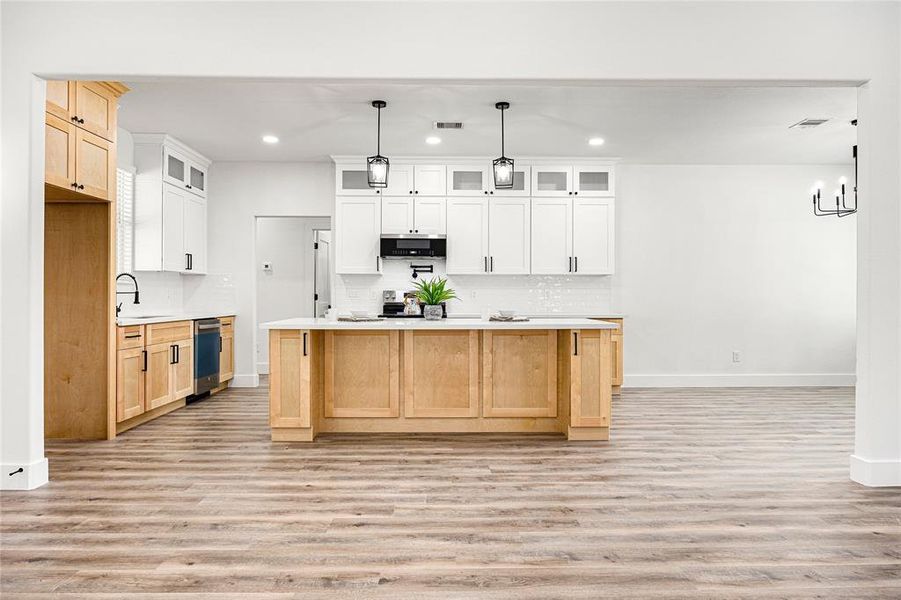 This photo showcases a modern, spacious kitchen with a large island featuring natural wood cabinetry and a white countertop. The kitchen includes white upper cabinets, a sleek black stove, and contemporary light fixtures, all set against a backdrop of light wood flooring, creating a bright and inviting space.