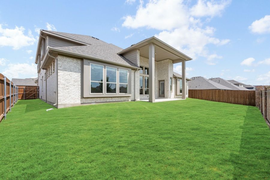 Rear exterior of two-story home with stone facade, covered patio, and fenced backyard with green lawn Rear exterior of two-story home with stone facade, covered patio, and fenced backyard with green lawn