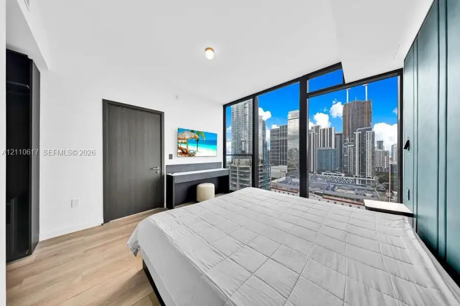Floor-to-ceiling windows featuring urban skyline scenery, wood-finish flooring, custom teal-paneled accent wall, and built-in dark-toned cabinetry Floor-to-ceiling windows featuring urban skyline scenery, wood-finish flooring, custom teal-paneled accent wall, and built-in dark-toned cabinetry