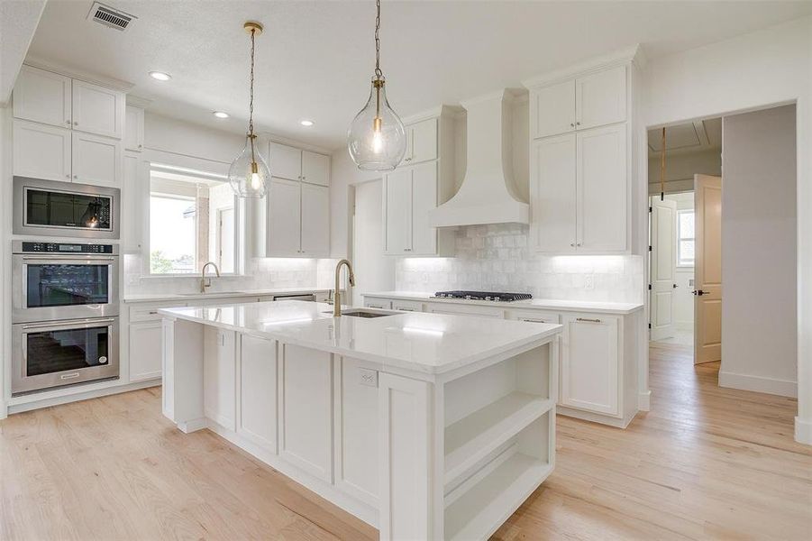 Kitchen featuring a sink, custom exhaust hood, appliances with stainless steel finishes, an island with sink, and light wood-style floors