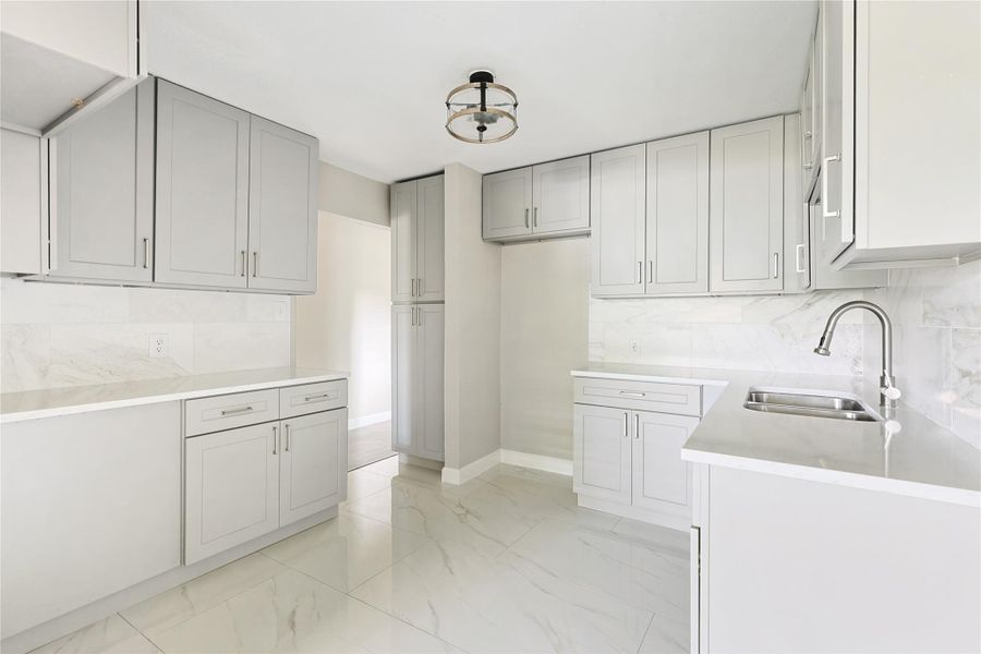 This kitchen features sleek gray cabinetry, modern fixtures, and a bright, open layout with a marble tile backsplash and flooring, creating a clean and contemporary feel.