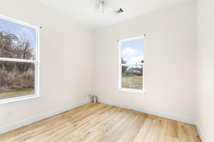 Bright second bedroom with light wood flooring, neutral walls, and two large windows offering a view of the outdoors.