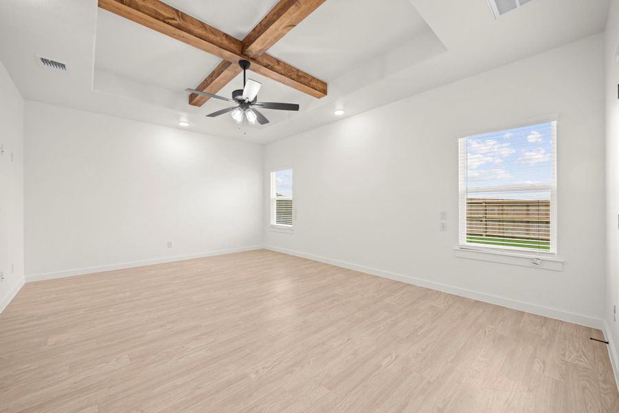 A spacious, naturally illuminated room features light flooring, a tray ceiling with wood beams, and a modern ceiling fan.