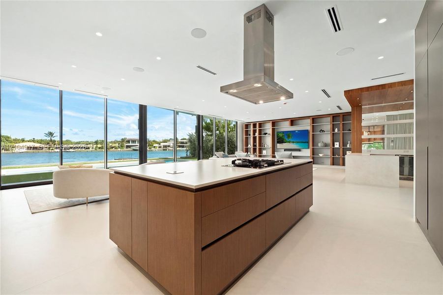 Expansive floor-to-ceiling windows, large kitchen island with cooktop, stainless steel range hood, integrated shelving, and light-toned flooring