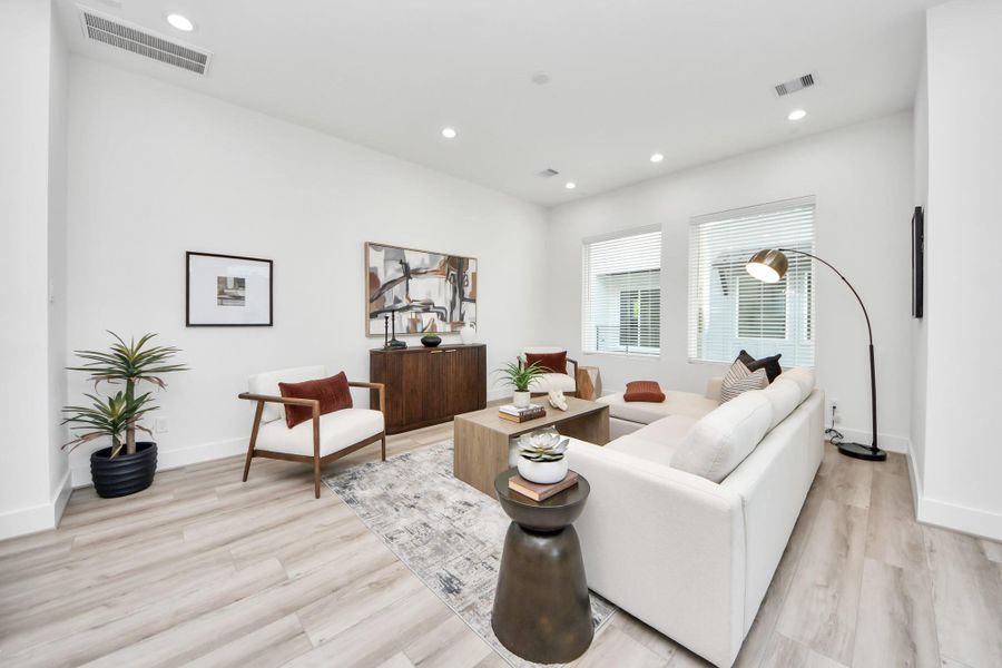 This is a bright, modern living room featuring light wood flooring, a white sectional sofa, and contemporary decor. It includes a stylish floor lamp, a sleek wooden cabinet, and large windows that let in plenty of natural light.